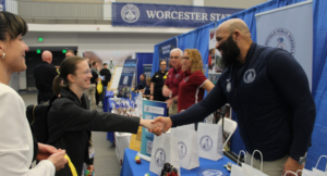 A woman shakes hands with a Worcester State University representative at a booth during an indoor event with several people and informational tables.