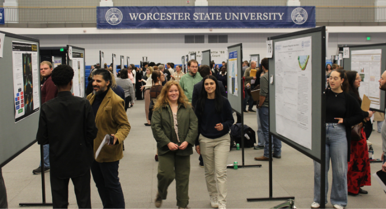 Students and attendees view and discuss research posters at a presentation event in a large hall at Worcester State University.