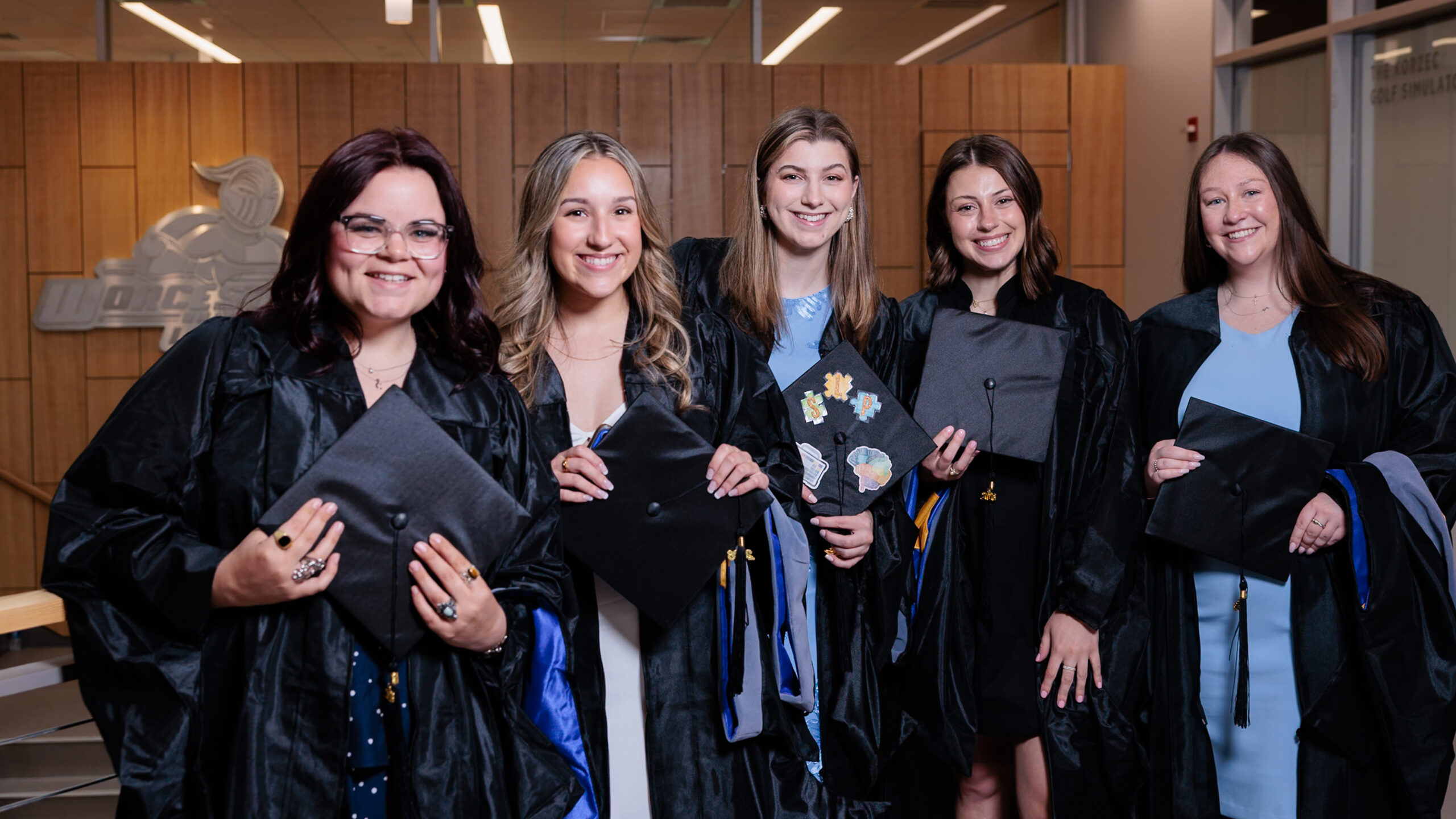 Five graduates in black robes stand indoors, holding decorated mortarboards and smiling at the camera.