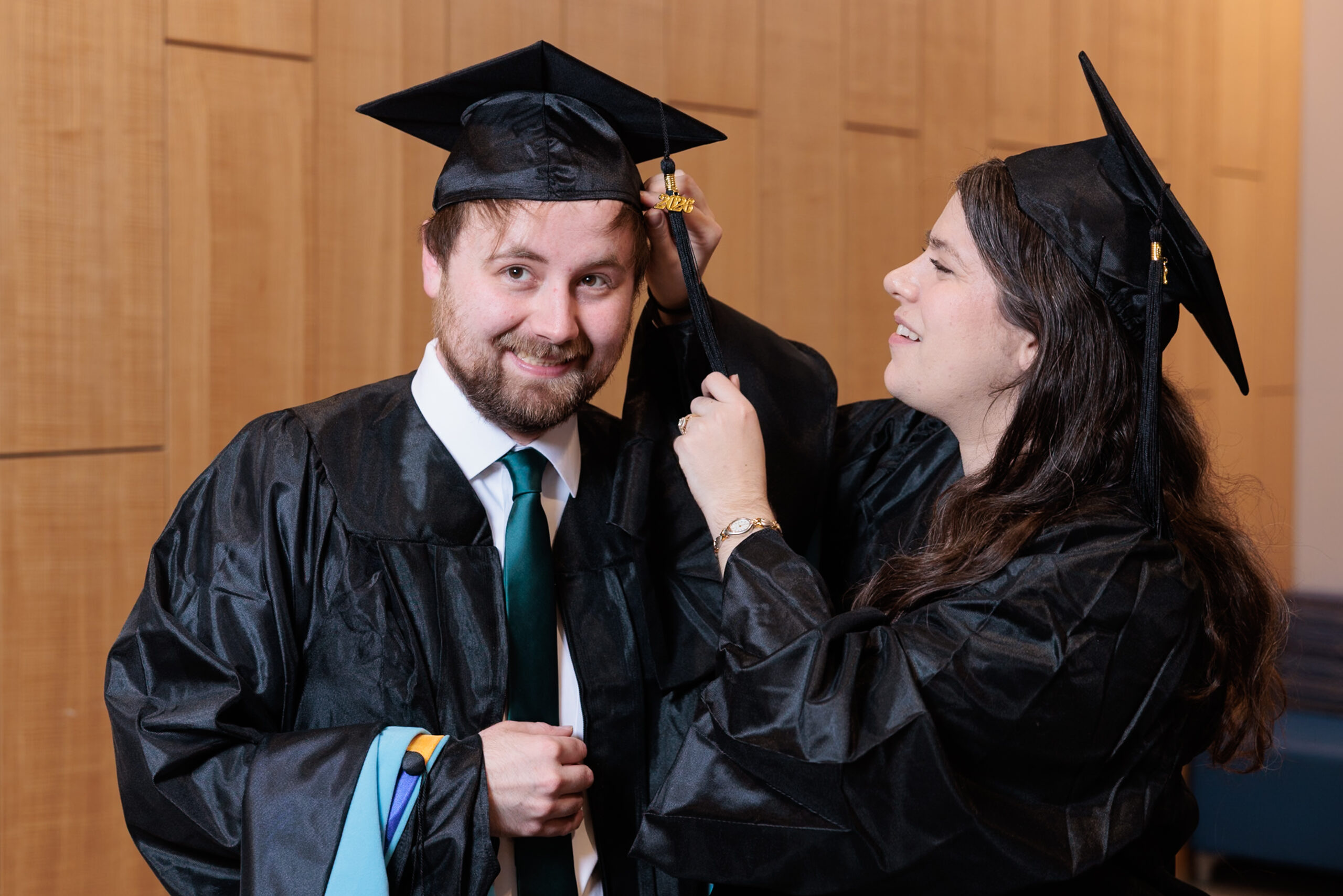 Two graduates in caps and gowns stand indoors; one smiles while the other adjusts the tassel on his graduation cap.
