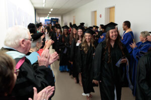 Graduates in caps and gowns walk down a hallway while faculty and others stand on either side clapping and celebrating their achievement.