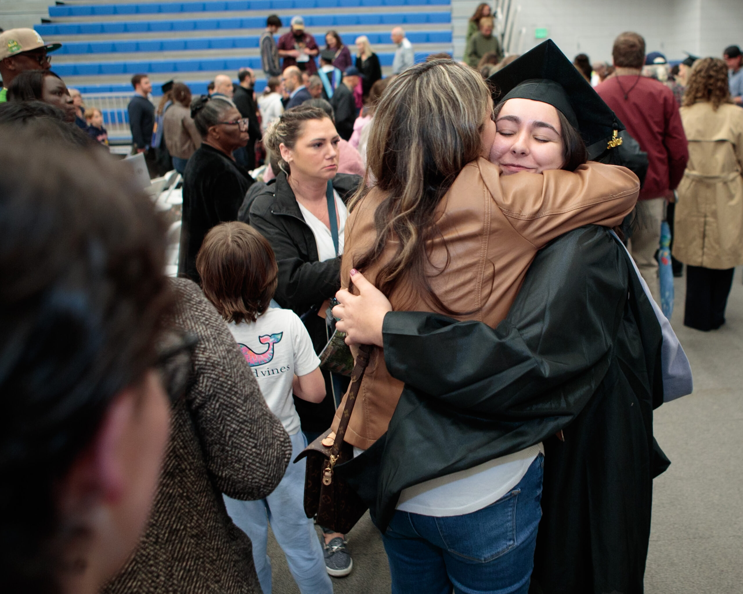 A graduate in cap and gown hugs a woman at a crowded indoor graduation ceremony, with people standing and talking in the background.