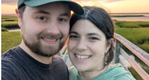 Two people smile for a selfie outdoors on a boardwalk with grassy fields and water in the background at sunset.