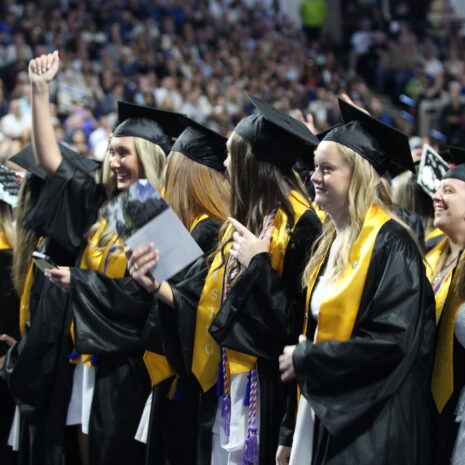 A group of graduates in caps and gowns stand and celebrate at a graduation ceremony, with one person raising a fist in the air.