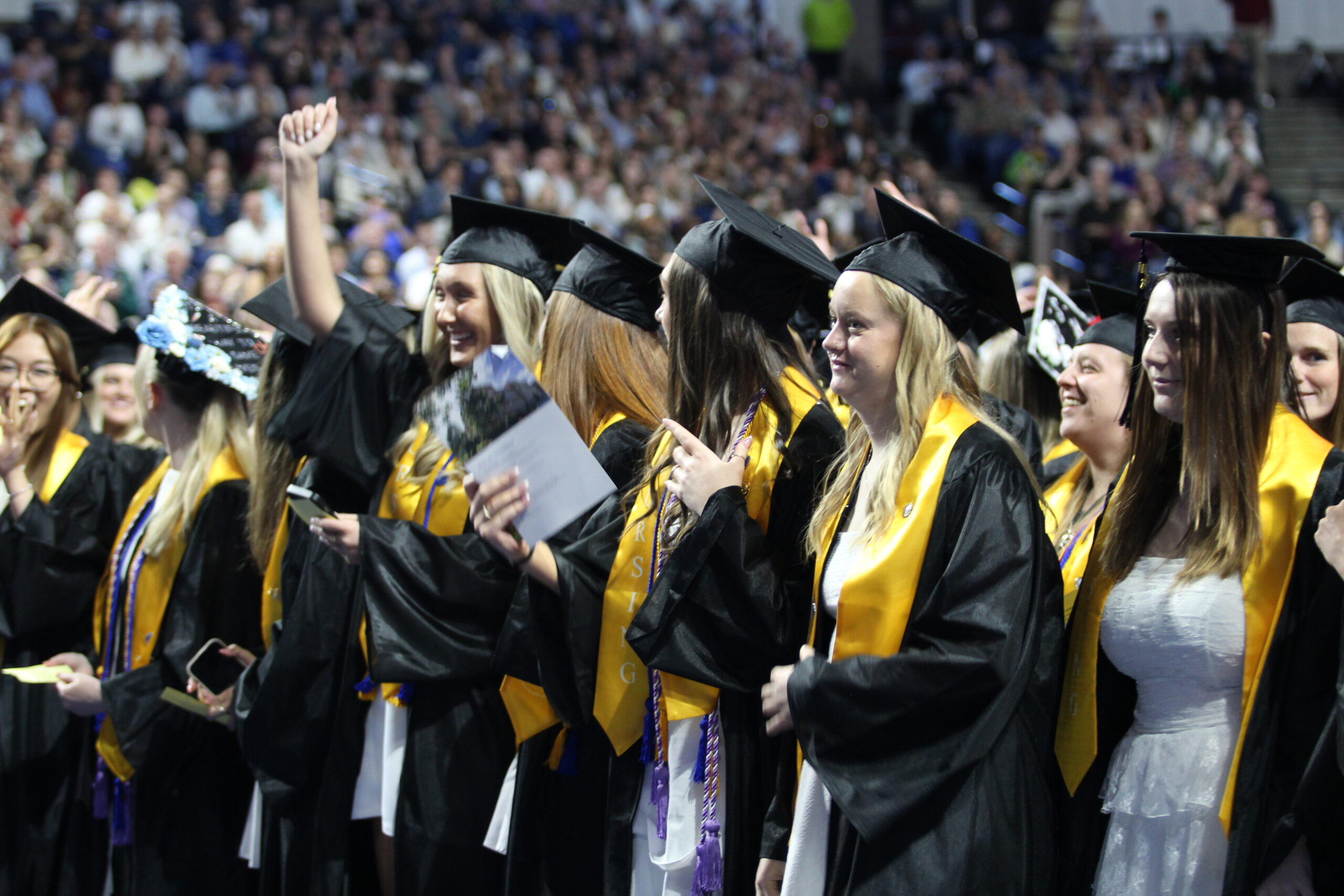 A group of graduates in caps and gowns stand and celebrate at a graduation ceremony, with one person raising a fist in the air.