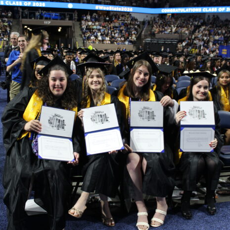 Four graduates in caps and gowns sit and hold diplomas, smiling at the camera during a graduation ceremony in a large arena.