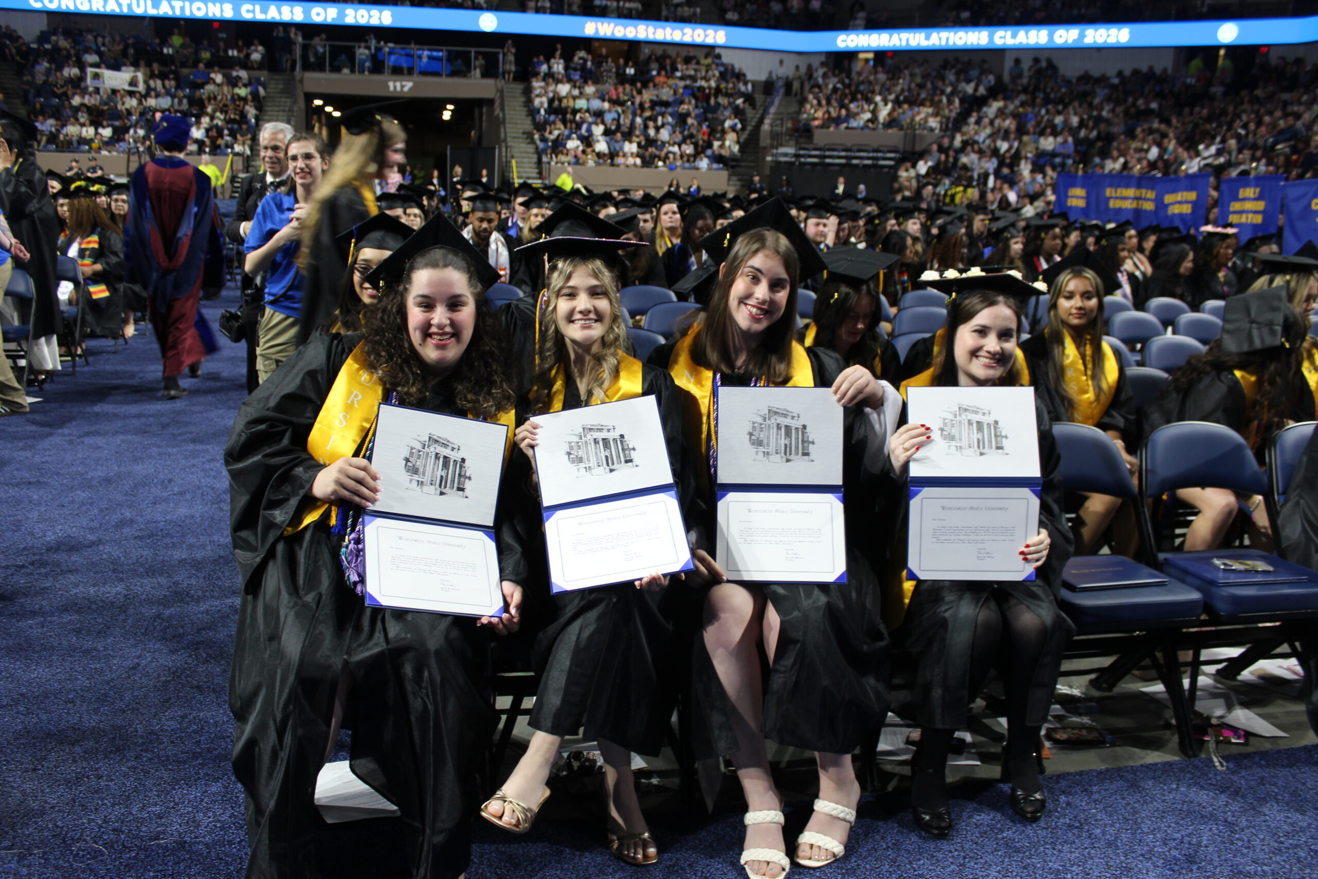 Four graduates in caps and gowns sit and hold diplomas, smiling at the camera during a graduation ceremony in a large arena.