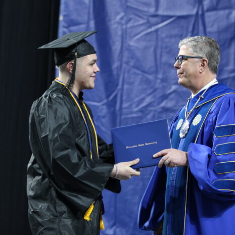 A graduate in a cap and gown receives a diploma from a man in academic regalia during a graduation ceremony.