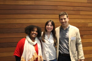 Three people stand side by side, smiling at the camera in front of a wooden panel wall. Two women are on the left and a man is on the right, all wearing name badges.
