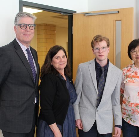 Four people stand indoors near a door; three are dressed in business attire, and one is wearing a floral blouse.