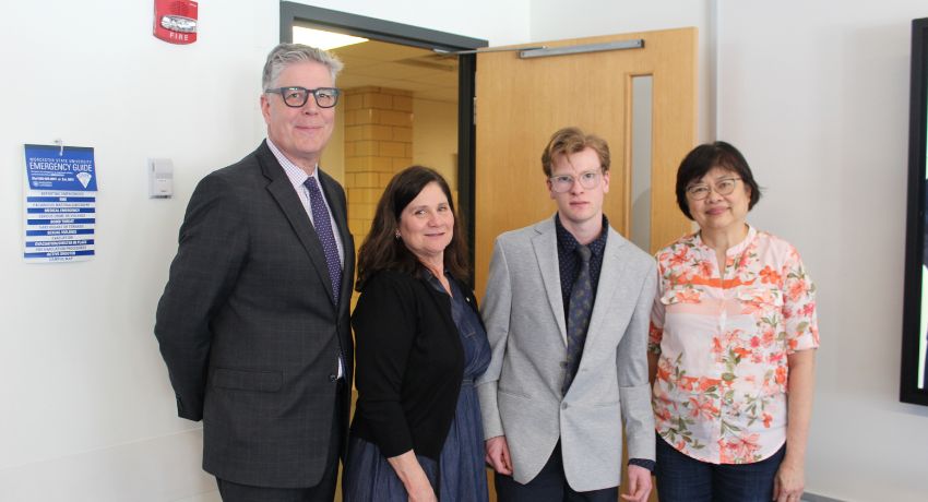 Four people stand indoors near a door; three are dressed in business attire, and one is wearing a floral blouse.