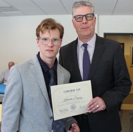 Two men pose indoors; one holds a certificate. People are seated at tables in the background.