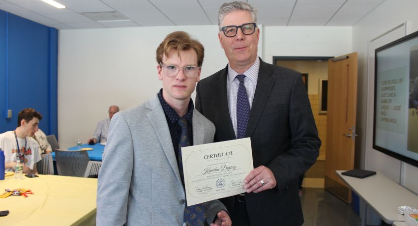 Two men pose indoors; one holds a certificate. People are seated at tables in the background.