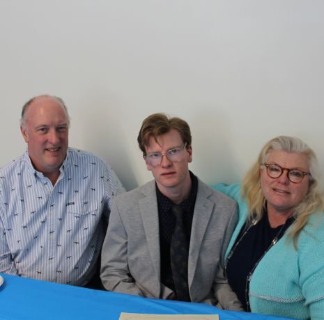 Three people sit at a table with a blue tablecloth, two water bottles, and a plate with food. The person in the center wears a suit; the two on either side are dressed casually.