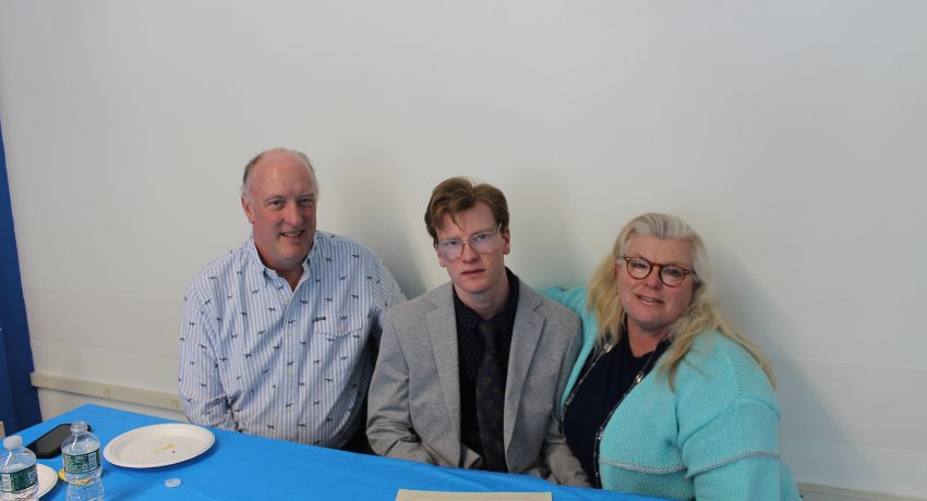 Three people sit at a table with a blue tablecloth, two water bottles, and a plate with food. The person in the center wears a suit; the two on either side are dressed casually.