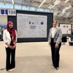 Two women stand in front of a large academic research poster titled "How Social Norms Shape Women’s Economic Participation in Pakistan" at a poster presentation event in a gymnasium.