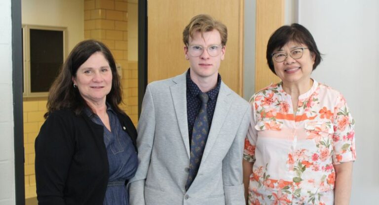 Three people stand indoors, posing for a photo; one man in a light suit and tie is between two women, one in a dark outfit and one in a floral top.