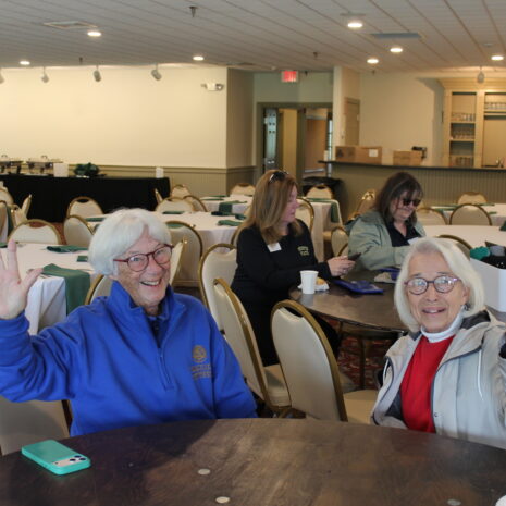Two older women sit at a round table, smiling and waving at the camera in a large, well-lit room with empty tables and chairs. Other people sit in the background.