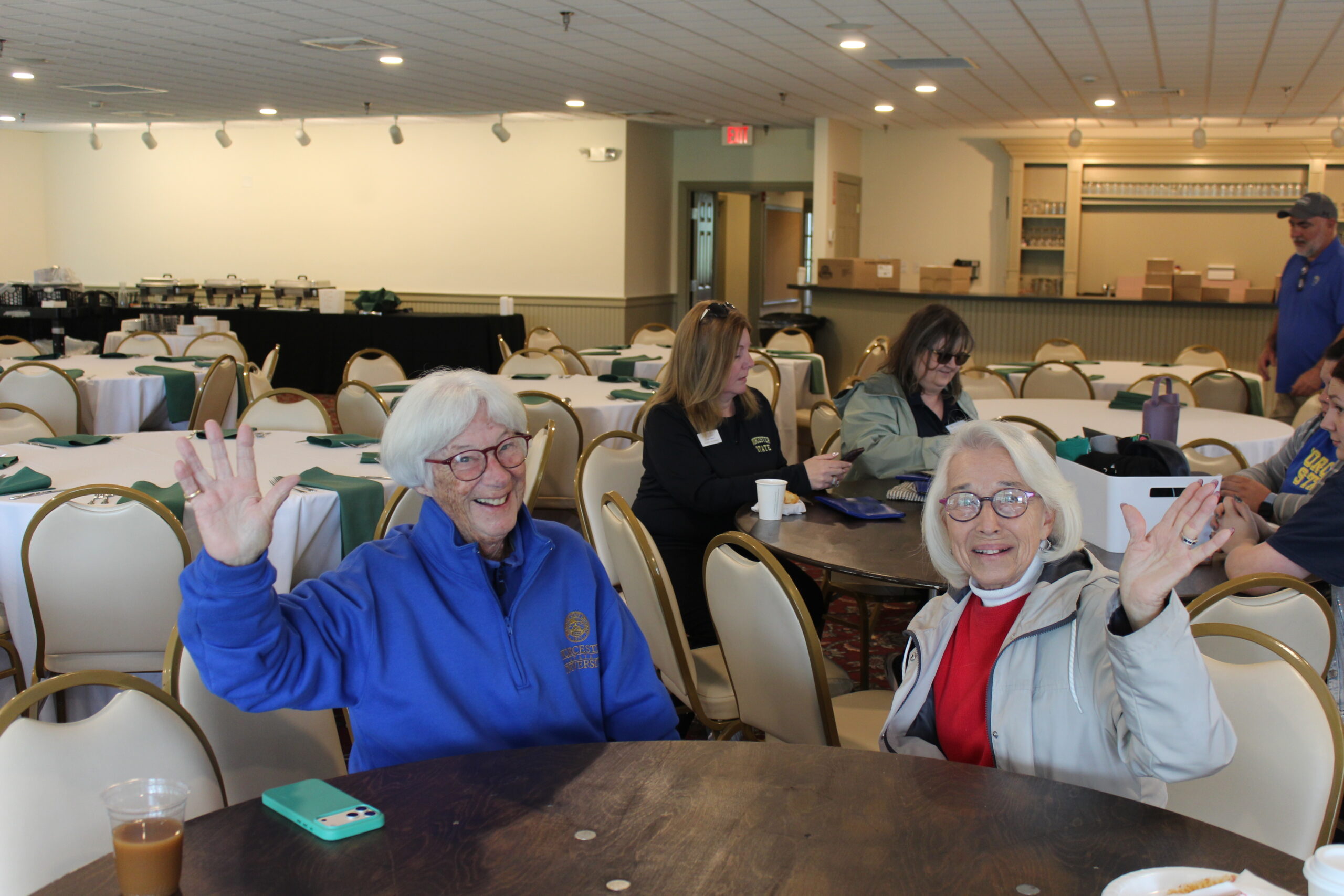 Two older women sit at a round table, smiling and waving at the camera in a large, well-lit room with empty tables and chairs. Other people sit in the background.