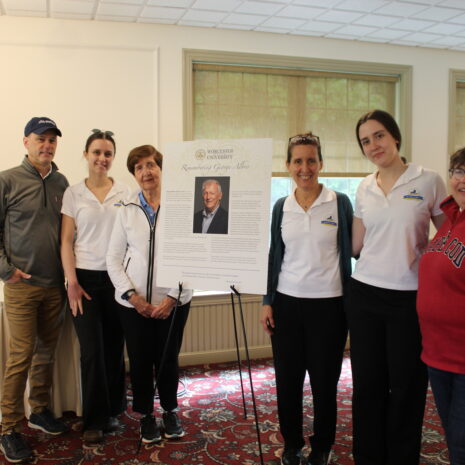 Six people stand indoors around a display board featuring a photo and biography of an older man; they are posing for a group photo on a patterned carpet.