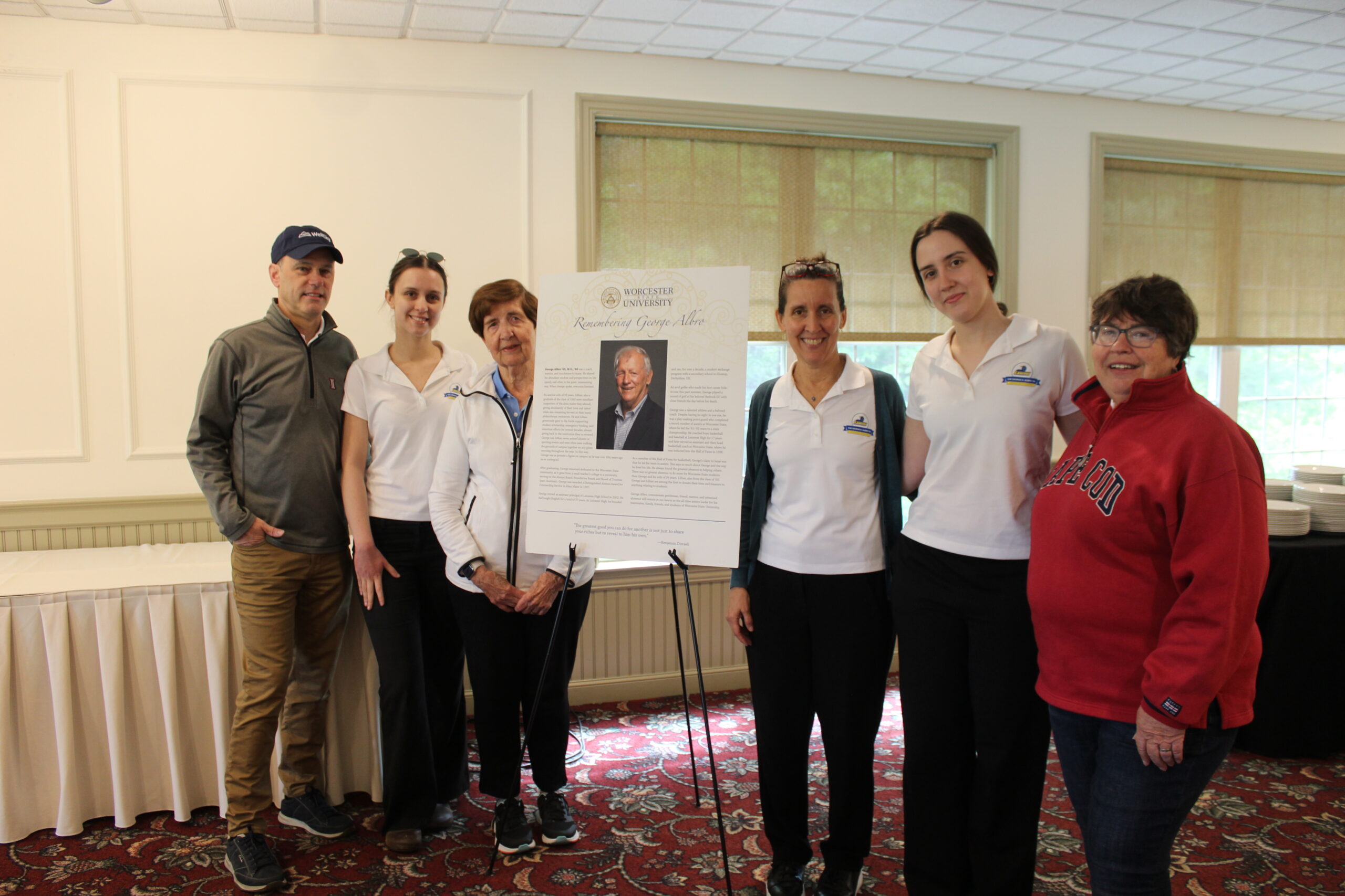 Six people stand indoors around a display board featuring a photo and biography of an older man; they are posing for a group photo on a patterned carpet.