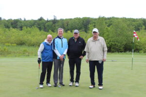 Four men stand on a golf green with clubs, posing for a photo near a flagstick, with trees and cloudy sky in the background.