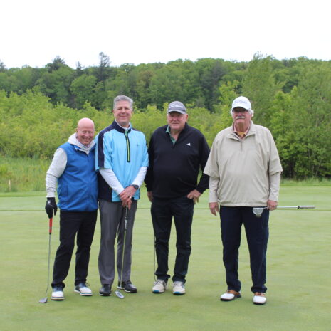Four men stand on a golf green with clubs, posing for a photo near a flagstick, with trees and cloudy sky in the background.