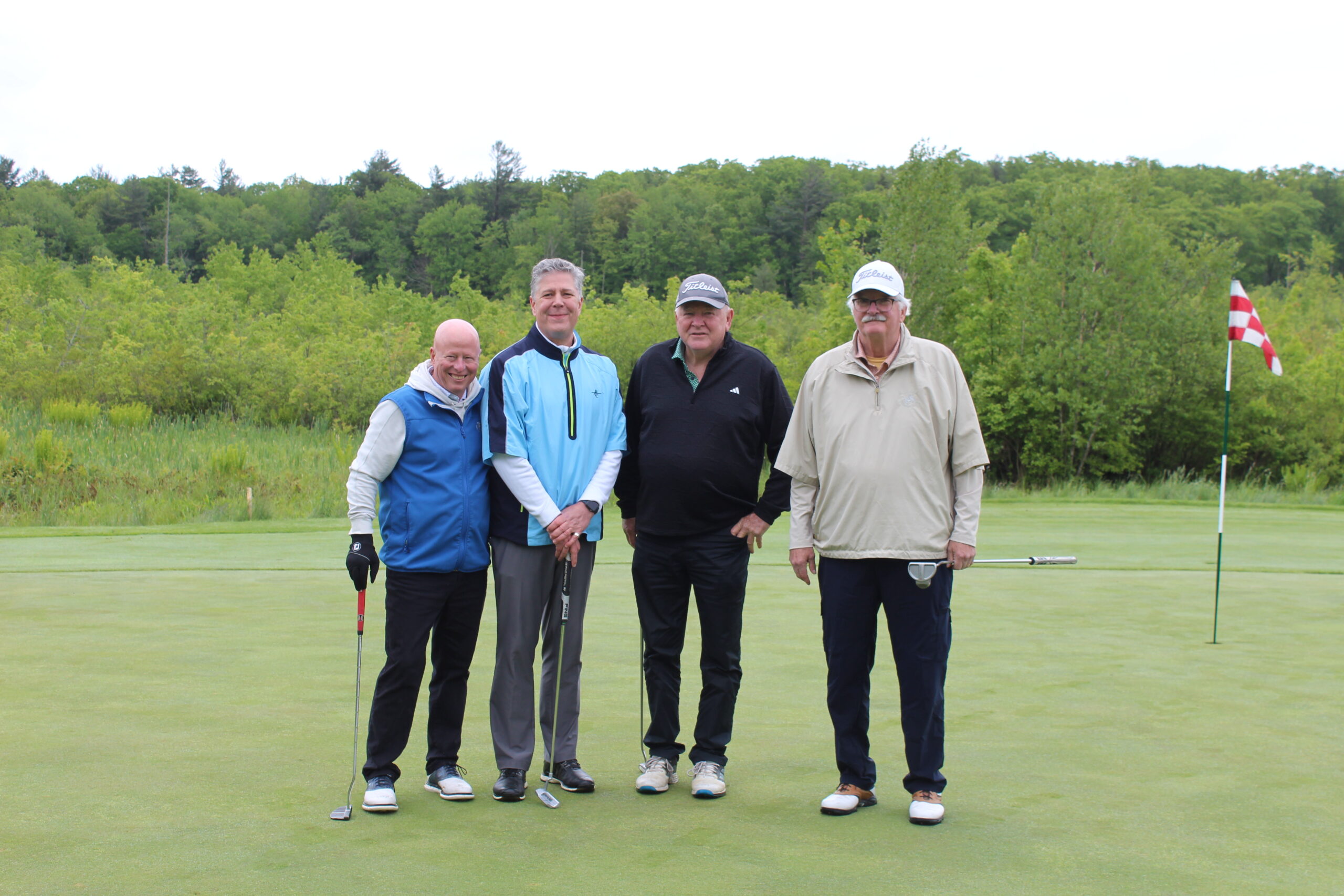 Four men stand on a golf green with clubs, posing for a photo near a flagstick, with trees and cloudy sky in the background.