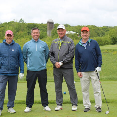 Four men in golf attire stand on a golf course with green grass, trees, and a silo in the background, posing for a group photo.
