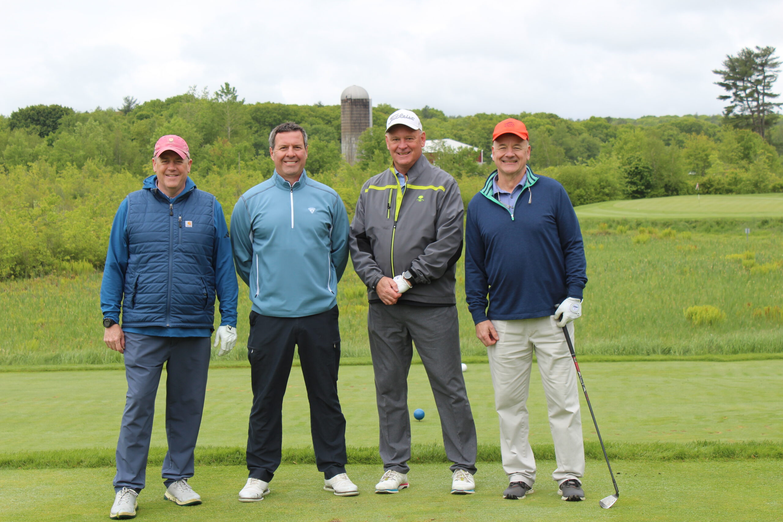 Four men in golf attire stand on a golf course with green grass, trees, and a silo in the background, posing for a group photo.