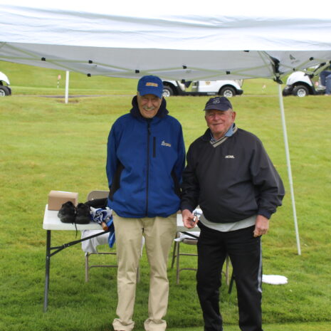 Two men stand and smile under a white canopy tent on a golf course, with golf carts and grassy hills in the background.