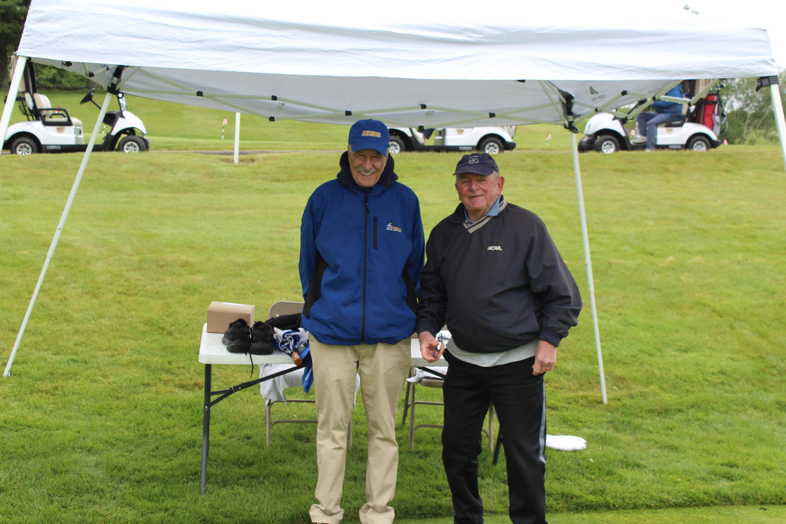 Two men stand and smile under a white canopy tent on a golf course, with golf carts and grassy hills in the background.