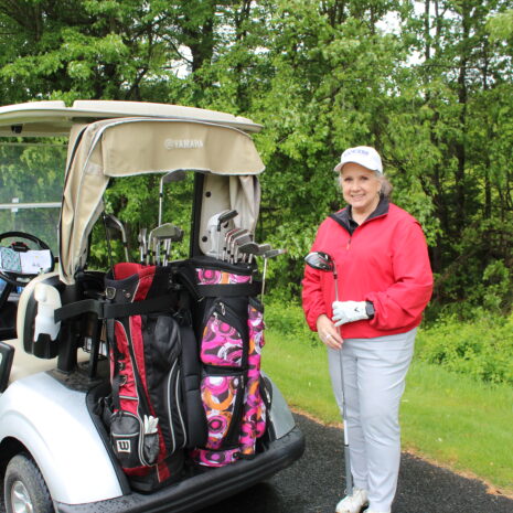 A woman in a red jacket stands next to a golf cart with golf bags on a paved path, surrounded by green trees.
