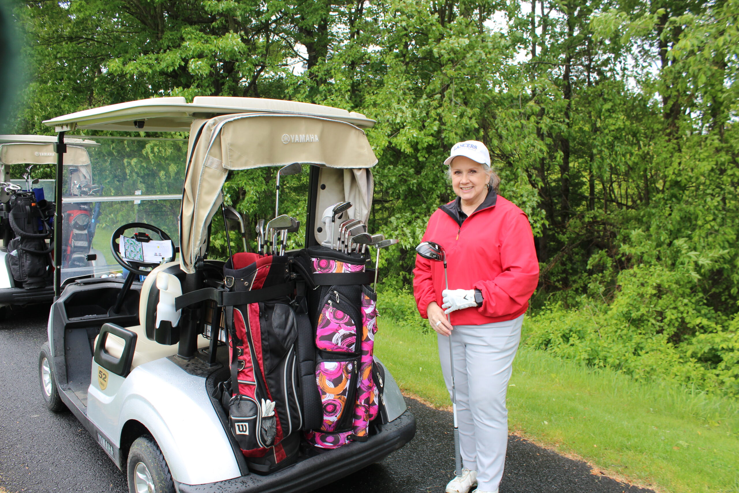 A woman in a red jacket stands next to a golf cart with golf bags on a paved path, surrounded by green trees.