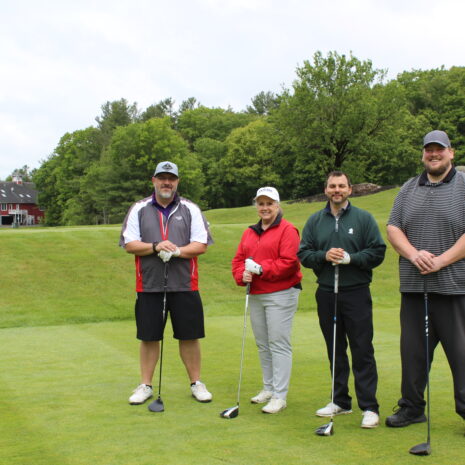 Four people stand on a golf course holding clubs, posing for a group photo with trees and a red house in the background.