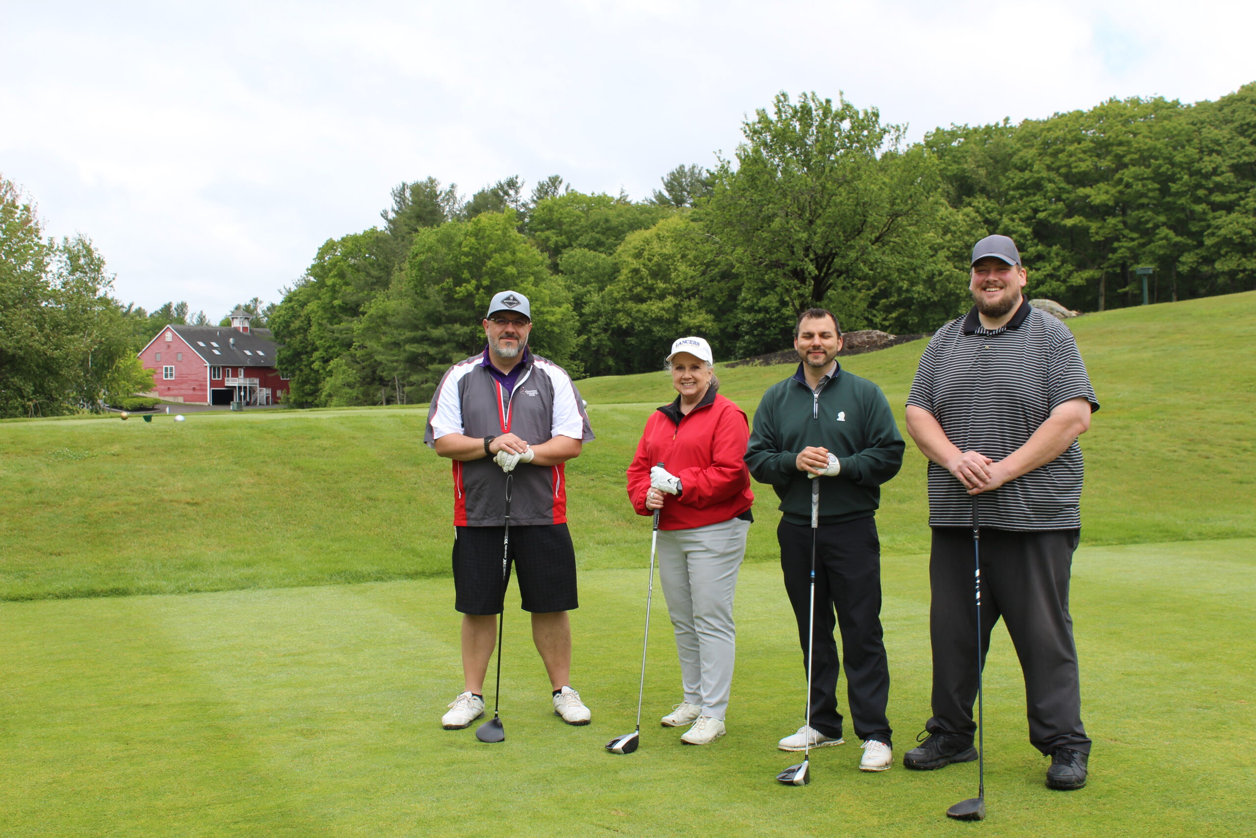 Four people stand on a golf course holding clubs, posing for a group photo with trees and a red house in the background.