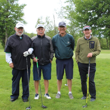 Four men stand on a golf course holding clubs, dressed in jackets and hats, with trees and grass visible in the background.