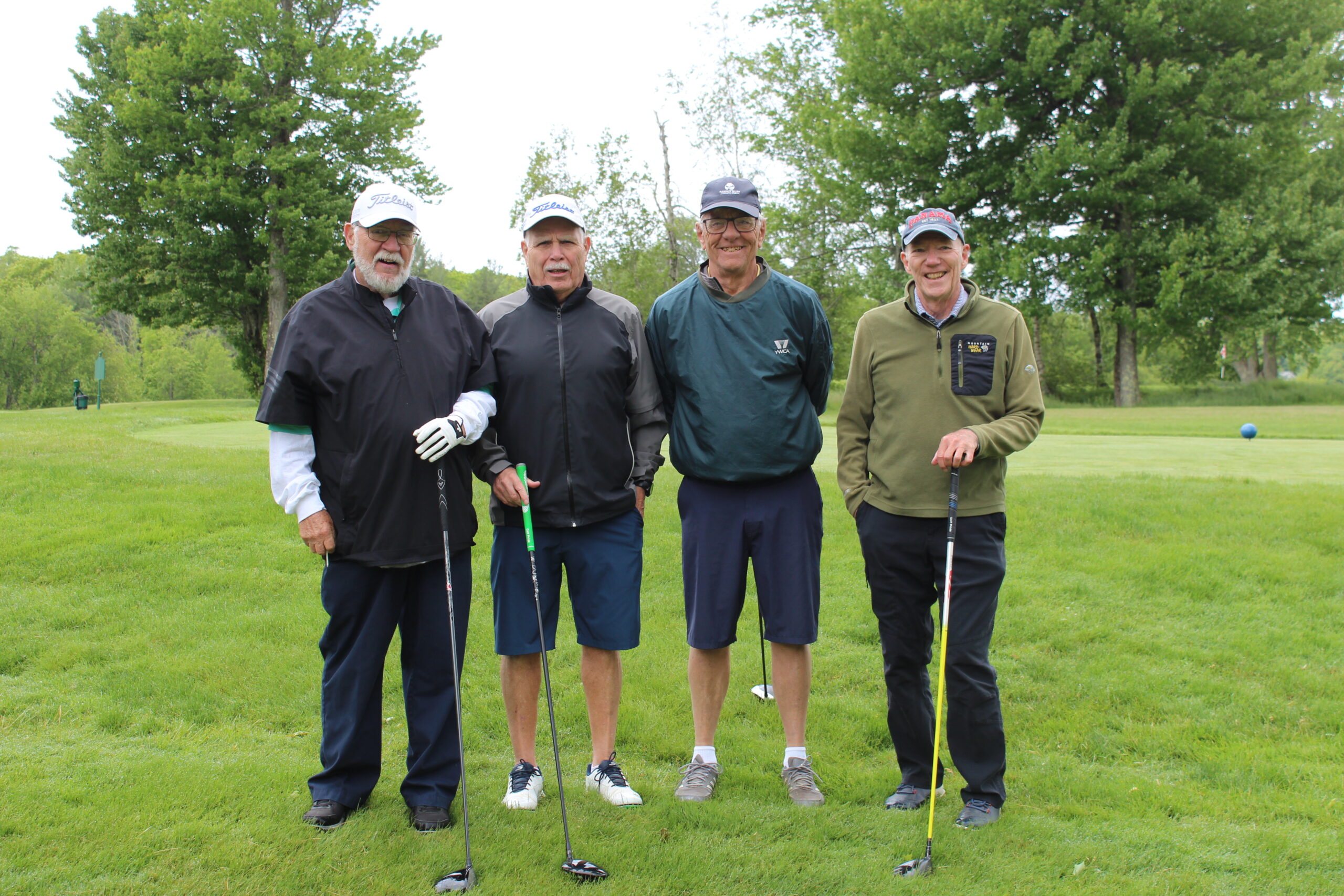 Four men stand on a golf course holding clubs, dressed in jackets and hats, with trees and grass visible in the background.