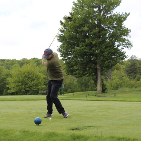 A person in a cap and jacket swings a golf club on a green golf course, with trees and bushes in the background.