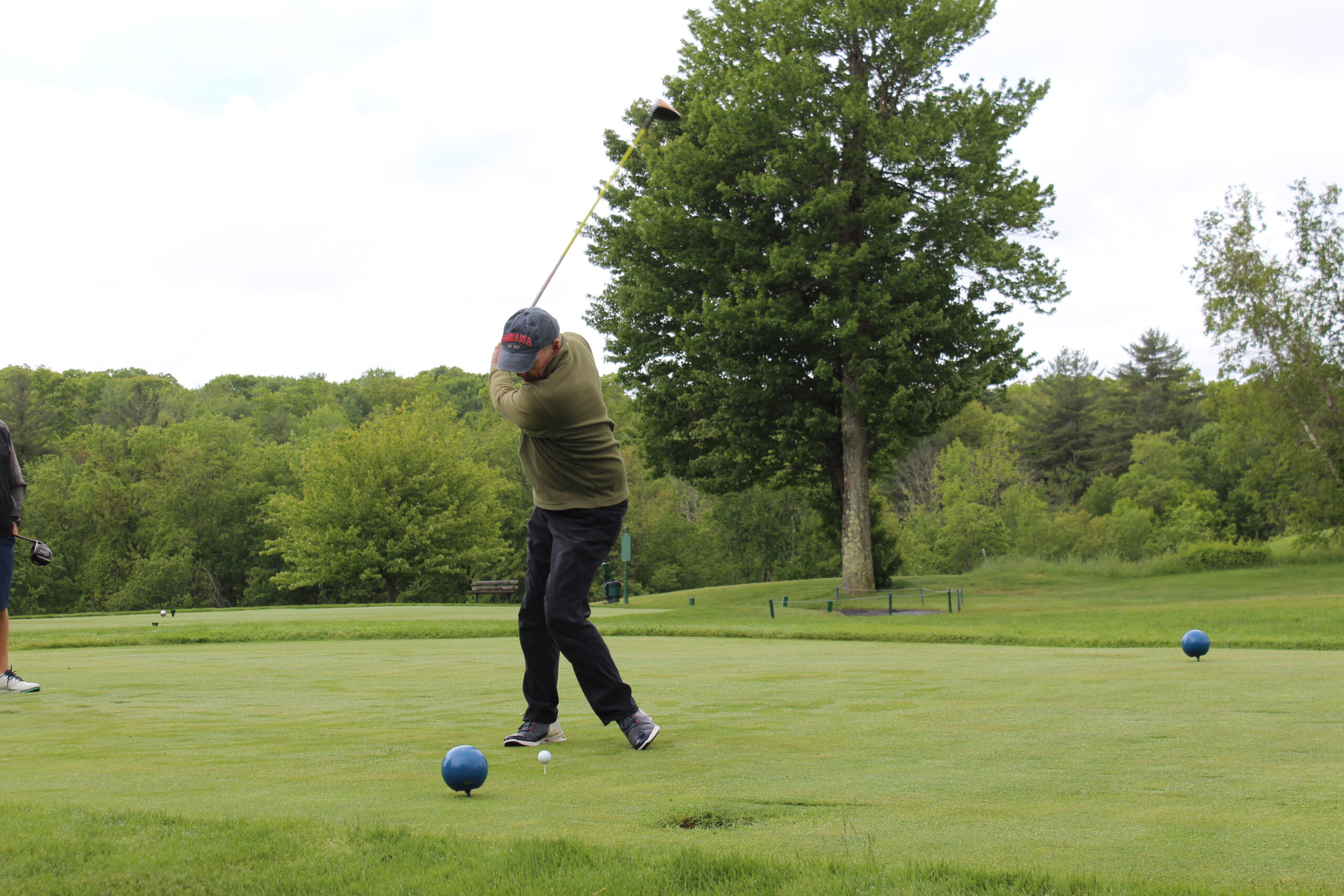 A person in a cap and jacket swings a golf club on a green golf course, with trees and bushes in the background.
