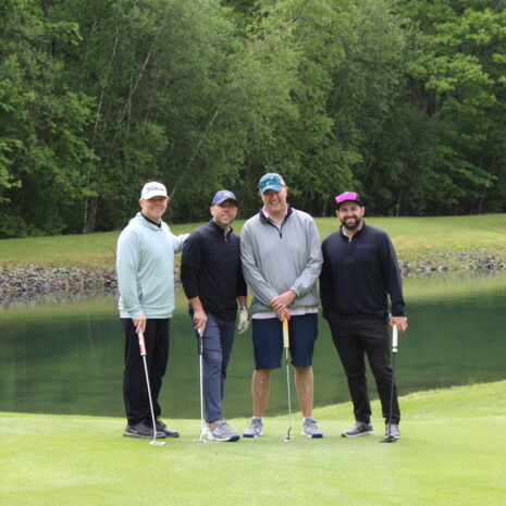 Four men standing on a golf course near a pond, each holding a golf club, with trees and greenery in the background.