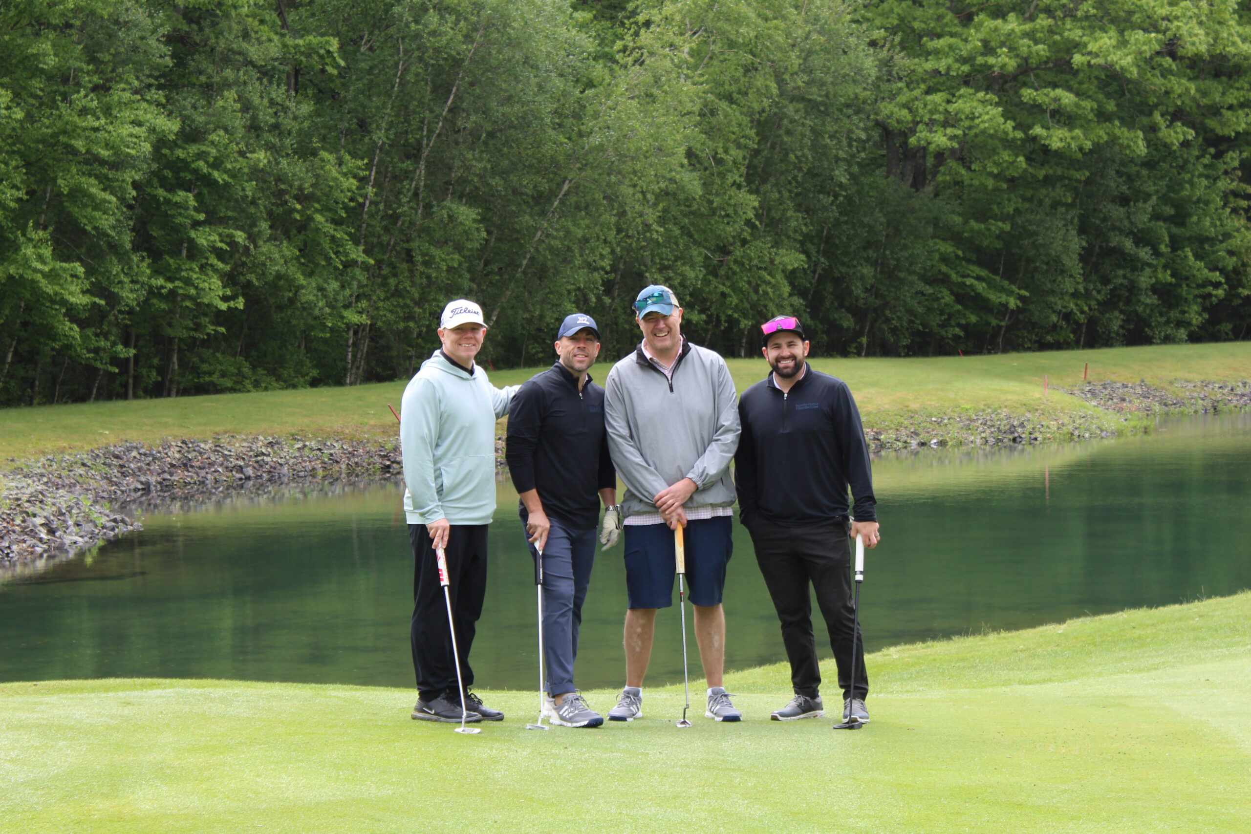 Four men standing on a golf course near a pond, each holding a golf club, with trees and greenery in the background.