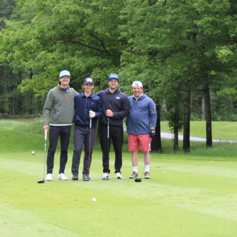 Four people stand together on a golf course, each holding a golf club, with trees and grass in the background.