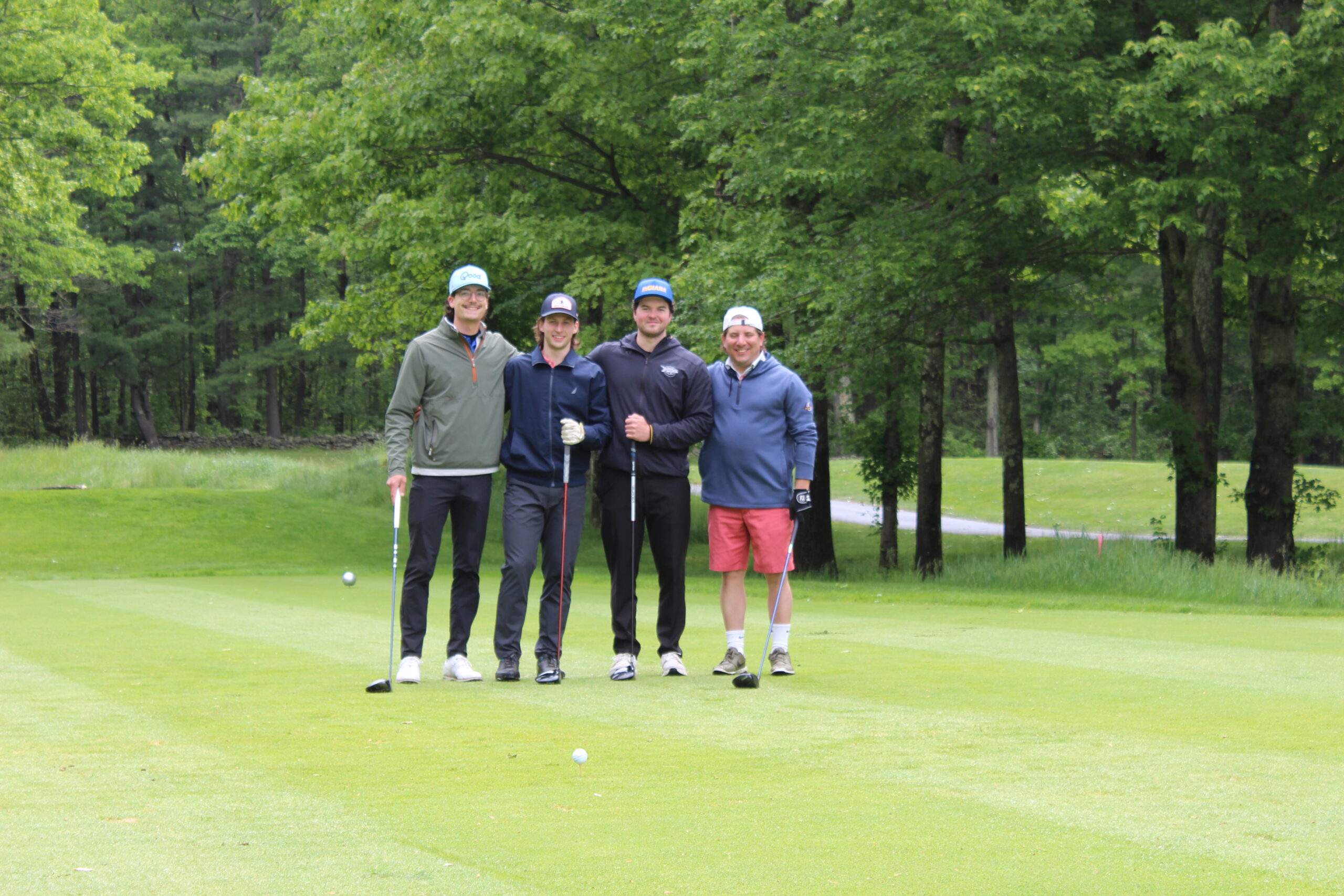 Four people stand together on a golf course, each holding a golf club, with trees and grass in the background.
