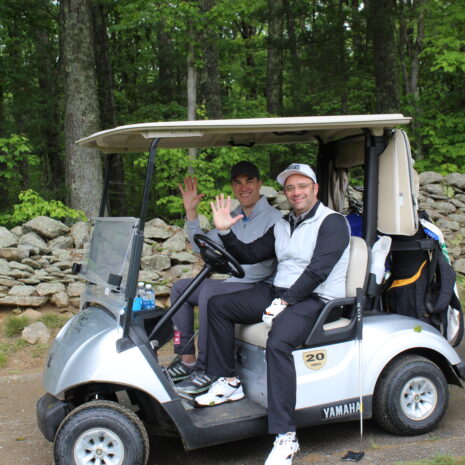 Two people sit in a golf cart on a dirt path, smiling and waving at the camera, with trees and a stone wall in the background.