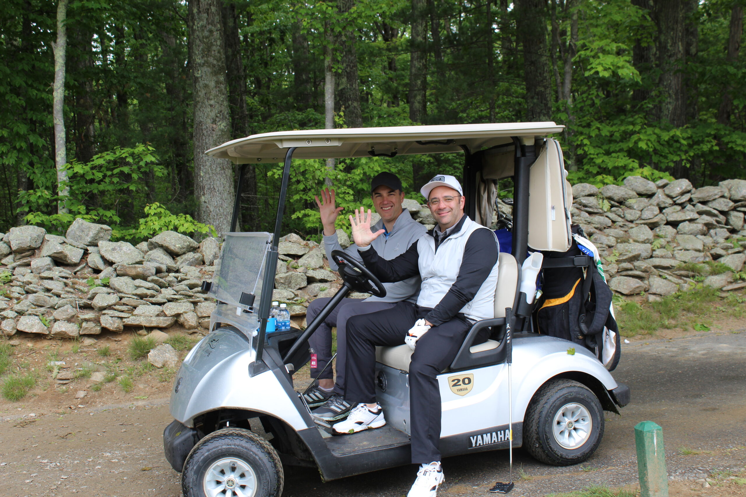 Two people sit in a golf cart on a dirt path, smiling and waving at the camera, with trees and a stone wall in the background.