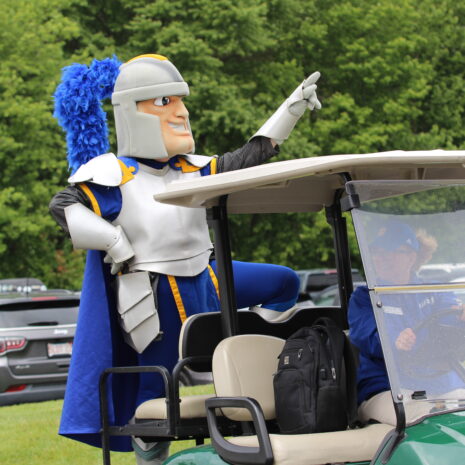 A person in a knight mascot costume stands with one foot on a golf cart, pointing ahead, while another person drives the cart; parked cars and trees are in the background.