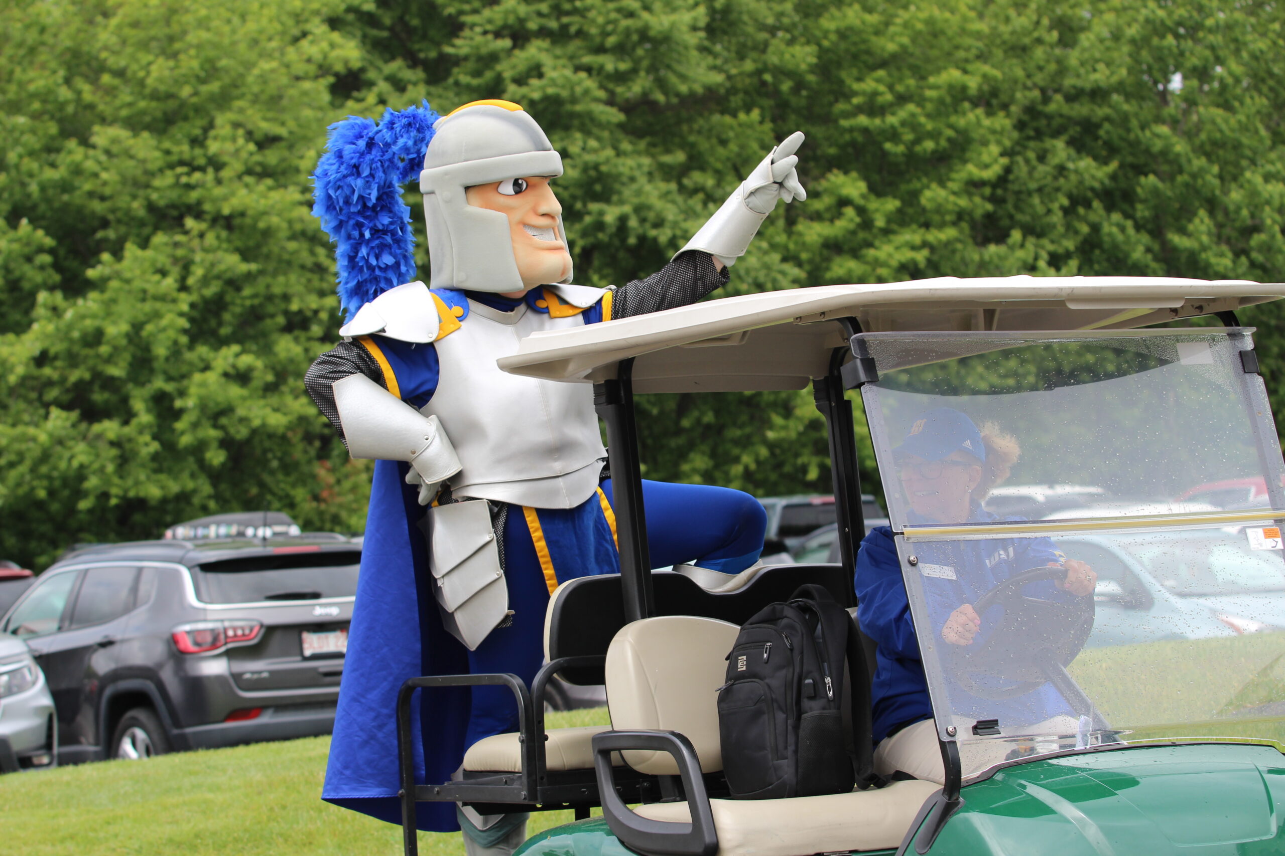 A person in a knight mascot costume stands with one foot on a golf cart, pointing ahead, while another person drives the cart; parked cars and trees are in the background.