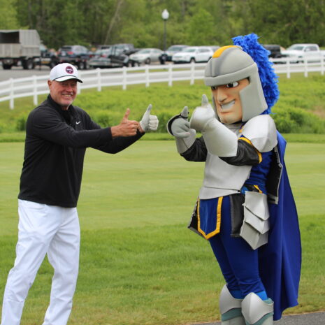 A man in golf attire and a person in a knight mascot costume stand on a golf course, both giving thumbs up.