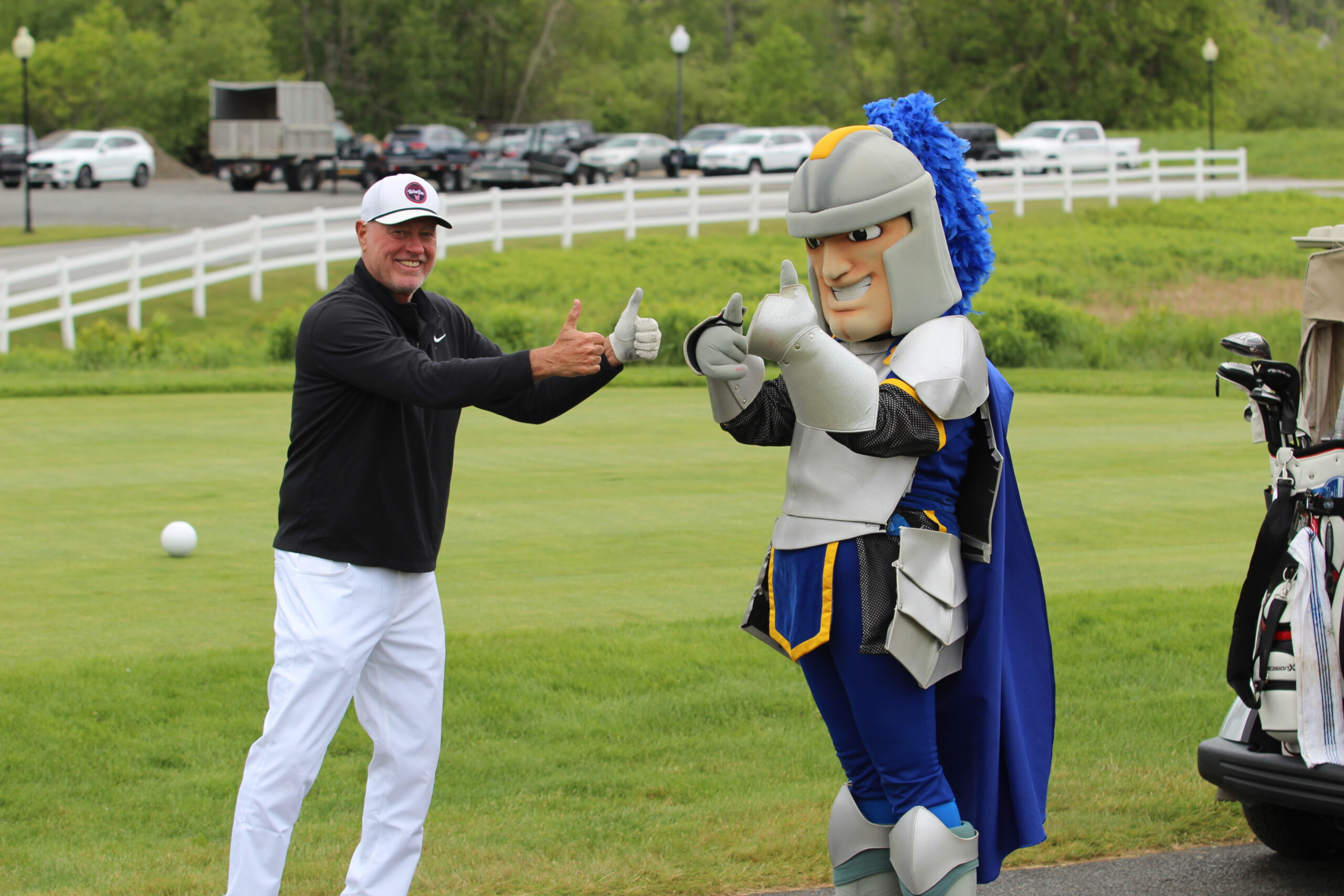 A man in golf attire and a person in a knight mascot costume stand on a golf course, both giving thumbs up.